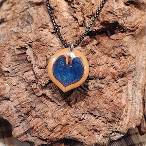 Op de afbeelding: Een ketting met een blauwe, hartvormige keramische hanger aan een zwarte ketting. De hanger is lichtbruin van kleur met een blauw hart in het midden. De ketting hangt aan een stuk drijfhout.