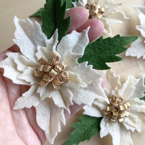 Può includere: Fiori di stella di Natale in feltro bianco con fiocchi di glitter dorati e foglie in feltro verde.