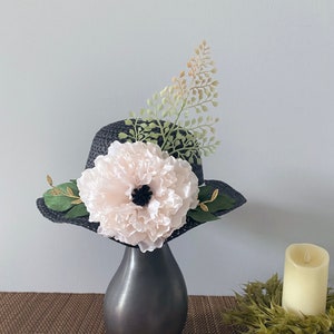 May include: A black straw hat with a large white flower and green foliage accents. The hat is sitting in a black vase on a brown woven mat.