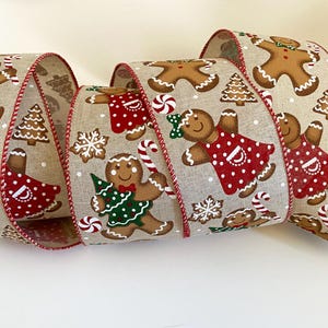 May include: A rolled-up ribbon featuring a repeating pattern of gingerbread people, Christmas trees, and snowflakes. The gingerbread figures wear red and green outfits and hold candy canes. The ribbon has a beige background with red trim.