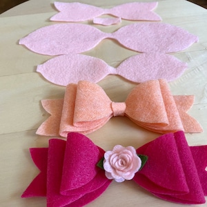 May include: A collection of felt bows in various stages of completion. The bows range in color from light pink to fuchsia, with one featuring a small pink flower and green leaves. The felt pieces are arranged on a light wooden surface.