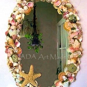 May include: An oval mirror framed with a dense arrangement of various seashells, including small spiral shells, bivalves, and sea urchins. A large, prominent tan starfish is attached to the lower left side. The text ADA Art Made is visible.