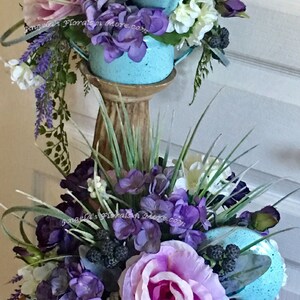 May include: Two blue watering cans with floral arrangements. The top watering can has white, purple, and pink flowers. The bottom watering can has purple, pink, and white flowers. Both watering cans have green leaves and berries.