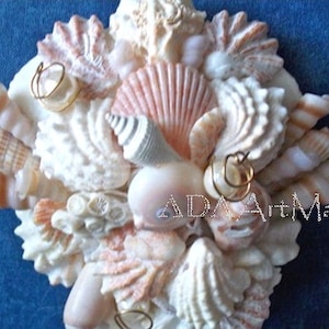 May include: A close-up of a seashell arrangement, featuring a variety of shapes, sizes, and colors. The shells are arranged in a circular pattern, with a few small pieces of wire or string woven through them. The shells are mostly white and pink, with some brown and orange accents.