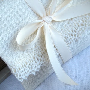 Könnte beinhalten: Ein weißes Leinenkissen mit weißer Spitzenborte und einem weißen Satinband, das zu einer Schleife gebunden ist.