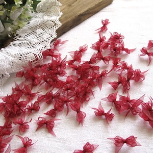 Könnte beinhalten: Eine große Menge kleiner, burgunderfarbener, durchsichtiger Schleifen aus Satinband. Die Schleifen sind auf einer weißen Oberfläche verstreut.