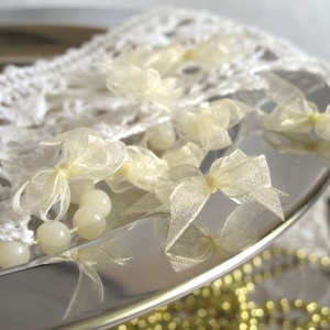 Peut inclure: Une collection de petits nœuds en ruban d'organza blanc cassé translucide disposés sur une surface argentée réfléchissante. L'ensemble comprend une bordure en dentelle crochetée blanche et un cordon de perles rondes et lisses.