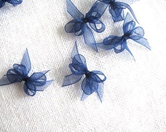 May include: A collection of small, navy blue, sheer ribbon bows. The bows are arranged on a white surface.