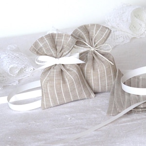 Può includere: Due piccole sacche di tessuto beige a righe legate con fiocchi di nastro bianco. Un nastro bianco e un quadrato di tessuto piegato sono accanto alle sacche. Tessuto di pizzo bianco sullo sfondo. Le sacche sono probabilmente per pot-pourri o bustine.
