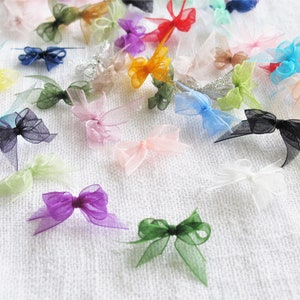 May include: Assortment of colorful sheer ribbon bows in various colors, including pink, blue, green, yellow, and black. The bows are arranged on a white fabric background.