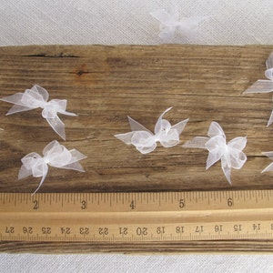 May include: Eight white tulle bows arranged on a wooden surface with a ruler for scale. The bows are approximately 1 inch in size.