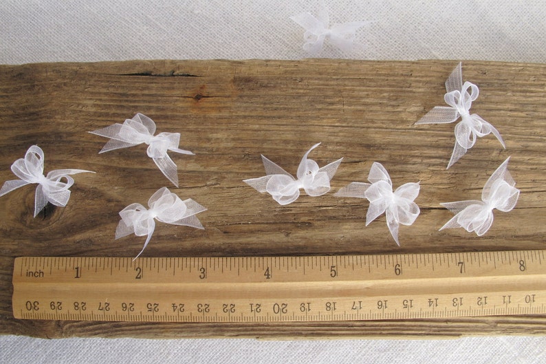May include: Eight white tulle bows arranged on a wooden surface with a ruler for scale. The bows are approximately 1 inch in size.