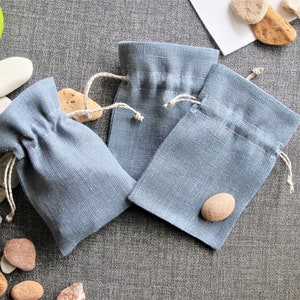 Könnte beinhalten: Drei kleine blaue Leinensäckchen mit weißen Kordeln. Die Säckchen liegen auf einer grauen, strukturierten Oberfläche mit kleinen Steinen, die um sie herum verstreut sind.