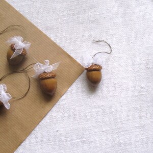 May include: Four brown acorns with white tulle bows tied around the tops. The acorns are strung on twine and are ready to be used as ornaments.