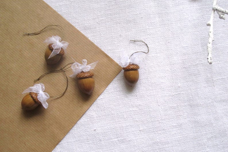 May include: Four brown acorns with white tulle bows tied around the tops. The acorns are strung on twine and are ready to be used as ornaments.