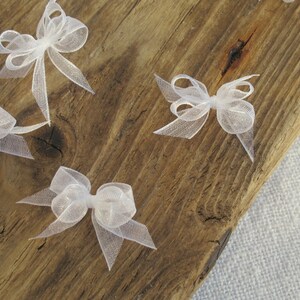 May include: Five white sheer ribbon bows on a wooden surface. The bows are tied in a simple knot and have a delicate, airy appearance.