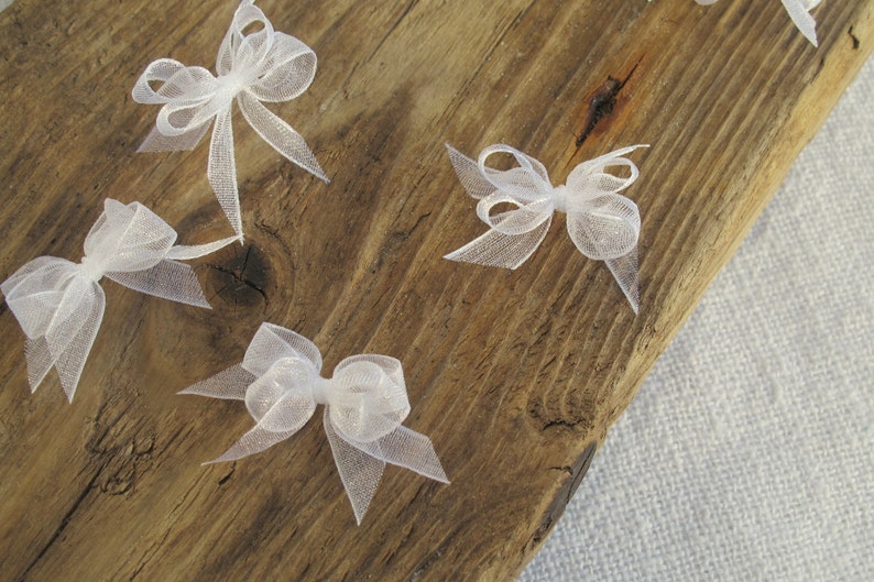 May include: Five white sheer ribbon bows on a wooden surface. The bows are tied in a simple knot and have a delicate, airy appearance.