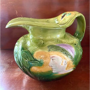 May include: A green ceramic pitcher with a peacock feather handle and a relief design of a woman's face. The pitcher has a green, brown, and yellow glaze.