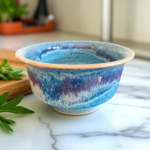 May include: A handmade ceramic bowl with a blue and purple glaze. The bowl has a light tan rim and a footed base. The interior of the bowl features a swirling pattern of blues and purples. The bowl is suitable for serving food.