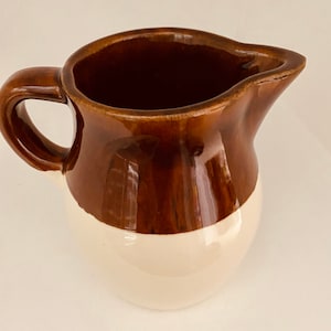May include: A two-toned ceramic pitcher with a handle and a spout. The top half is a glossy, dark brown, while the bottom half is a creamy white. The pitcher has a classic, rounded shape, suitable for serving beverages.