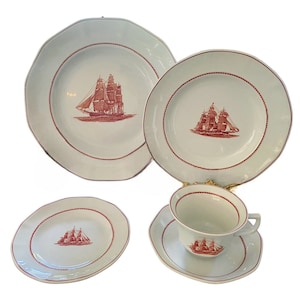 May include: A set of white porcelain dinnerware with a red ship design. The set includes a large dinner plate, a smaller dinner plate, a saucer, and a teacup.