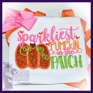 May include: A white shirt featuring an embroidered design with a glittery orange pumpkin, green vines, and the text "sparkliest PUMPKIN in the PATCH" in pink, orange, and green. A large pink hair bow is attached to the top left corner of the shirt.