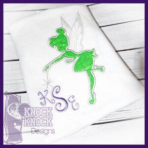 May include: A white t-shirt featuring an embroidered green glitter fairy silhouette with silver wings and a wand. Below the fairy, the letters KSE are embroidered in a purple cursive font. The logo KNOCK KNOCK DESIGNS is visible in the corner.