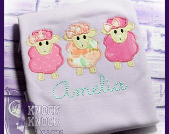 May include: A lavender t-shirt with three embroidered sheep wearing pink, floral, and white hats. The name "Amelia" is embroidered in green below the sheep. The text "Knock Knock Designs" is in the bottom left corner.