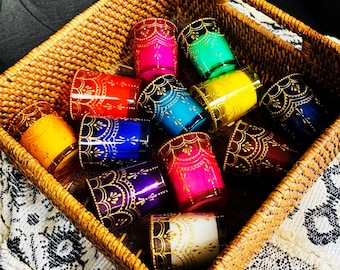 Puede incluir: Una colección de doce portavelas cilíndricos de vidrio en varios colores vibrantes, incluyendo rojo, amarillo, azul, verde, morado y blanco. Cada vaso presenta un patrón de filigrana pintado en oro a lo largo del borde superior.