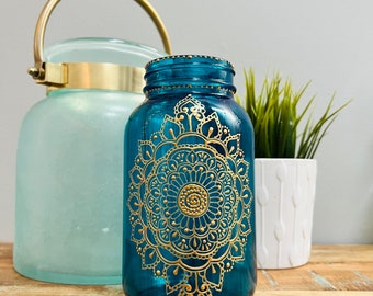 May include: Teal blue glass jar with a gold mandala design. The jar is sitting on a wooden surface.