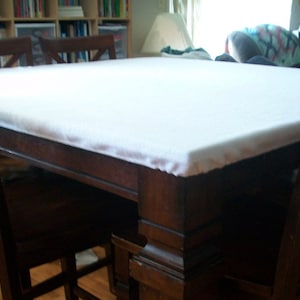 May include: A dark wood dining table with a white tablecloth. The table has a rectangular shape and is surrounded by four chairs. The chairs are also dark wood and have a simple design.