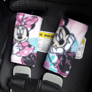 May include: Two pink and blue car seat shoulder strap covers with a Minnie Mouse design. The covers are made of soft fleece fabric and feature a pink polka dot pattern. The covers are attached to the car seat straps with velcro.
