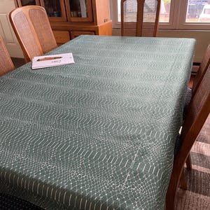 May include: A rectangular, green tablecloth with a white, snake-skin pattern covers a wooden dining table. The table is surrounded by wooden chairs with woven backs. A notepad and pen rest on the table.