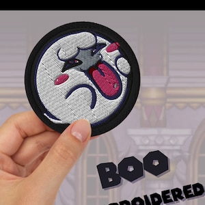 May include: A hand is holding a black and white embroidered patch of a ghost with a pink tongue sticking out. The patch says "BOO EMBROIDERED PATCH" in black text.
