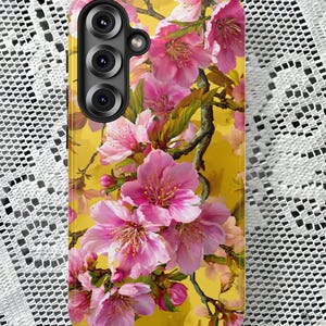 May include: A phone case featuring a vibrant floral design. Pink cherry blossoms with green leaves and brown branches are set against a yellow background. The case has a camera module with three lenses.