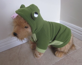 May include: A small brown dog wearing a green frog costume with a pink tongue sticking out. The costume has a hood with large white eyes and a green body with a light green trim.