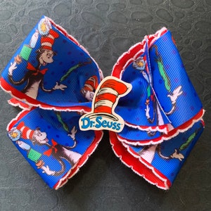 May include: A blue and red hair bow with a white scalloped edge. The bow features a repeating pattern of the Cat in the Hat from Dr. Seuss. The center of the bow has a Dr. Seuss logo.