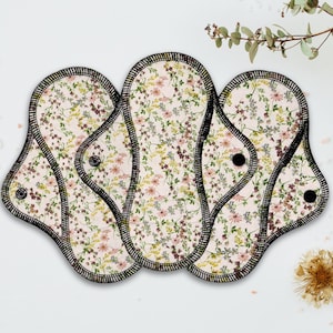 May include: Reusable cloth menstrual pads with a floral pattern in pink, yellow, and purple on a cream background. The pads have a black stitched border and black snaps. The pads are arranged on a white surface.