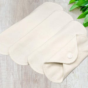 May include: Three off-white, reusable cloth menstrual pads. One pad is folded and secured with a white snap closure. The pads are arranged on a light wood-grain surface, with green leaves in the background.