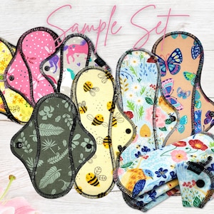 May include: A collection of reusable cloth menstrual pads with various colorful patterns, including floral, butterflies, bees, and a unicorn. The pads have a black trim and snap closures. The words "Sample Set" are written in pink script.
