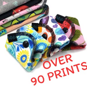 May include: A collection of reusable cloth pads in various colorful patterns, including floral, hearts, and polka dots. Each pad has a black snap closure and black stitching. The text "OVER 90 PRINTS!" is displayed in red.