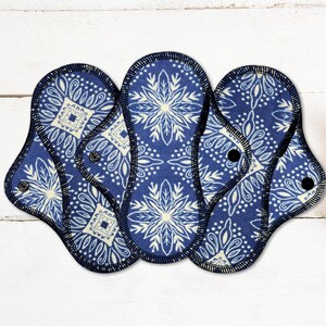May include: Three reusable cloth menstrual pads with a blue and white floral pattern. Each pad has a dark blue border and a snap closure. The pads are arranged on a white wooden surface.