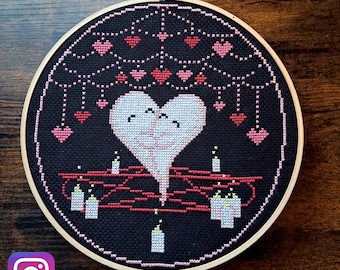 Könnte beinhalten: Ein schwarzer Stickrahmen mit einem Kreuzstich-Design eines weißen Herzens mit zwei Gesichtern, umgeben von rosa Herzen und Kerzen. Der Text "@little_stitchtx" befindet sich am unteren Rand des Rahmens.