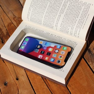 May include: A black smartphone is nestled within a carved-out section of an open book. The phone's screen shows app icons. The book has brown covers and visible pages. The scene is set on a wooden surface. The text "THE RETURN THE KING" is visible.