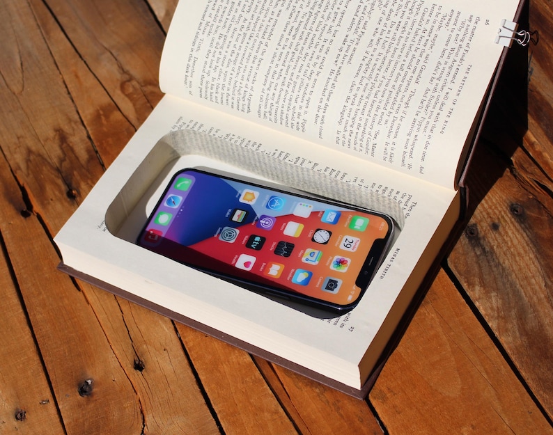 May include: A black smartphone is nestled within a carved-out section of an open book. The phone's screen shows app icons. The book has brown covers and visible pages. The scene is set on a wooden surface. The text "THE RETURN THE KING" is visible.