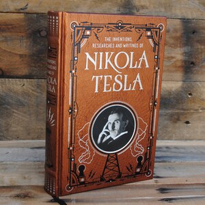 May include: A brown leather-bound book with gold accents and a portrait of Nikola Tesla on the cover. The title reads "The Inventions, Researches and Writings of Nikola Tesla".