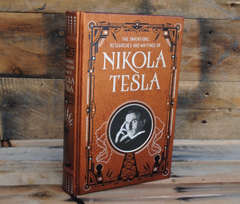 May include: A brown leather-bound book with gold accents and a portrait of Nikola Tesla on the cover. The title reads "The Inventions, Researches and Writings of Nikola Tesla".
