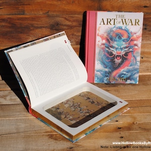 Puede incluir: Un libro hueco con una cubierta roja y el título "El Arte de la Guerra" impreso en blanco. El libro está abierto para revelar un compartimento oculto en el interior. El compartimento está forrado con papel blanco y tiene una pequeña abertura rectangular que revela una imagen colorida de un dragón.