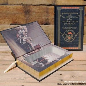 Può includere: Un libro cavo blu navy intitolato "The Constitution of the United States of America" con un vano nascosto. Il libro ha lettere dorate e bordi delle pagine dorati. Il libro aperto rivela un'illustrazione interna. Un segnalibro color crema è visibile.