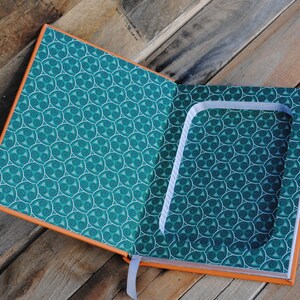 Puede incluir: Un libro abierto con un compartimento oculto en el interior. El libro tiene una cubierta de color turquesa con un patr&oacute;n de espiral y un borde naranja. El compartimento est&aacute; forrado con tela blanca.